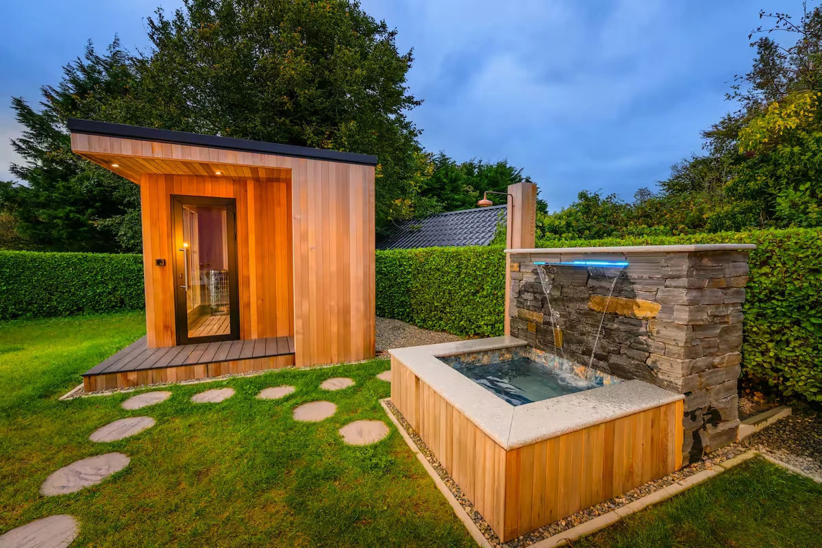 An image of Lily's Pink House Sauna and Plunge Pool with stepping stones leading to each of them on a calm evening with blue skies.