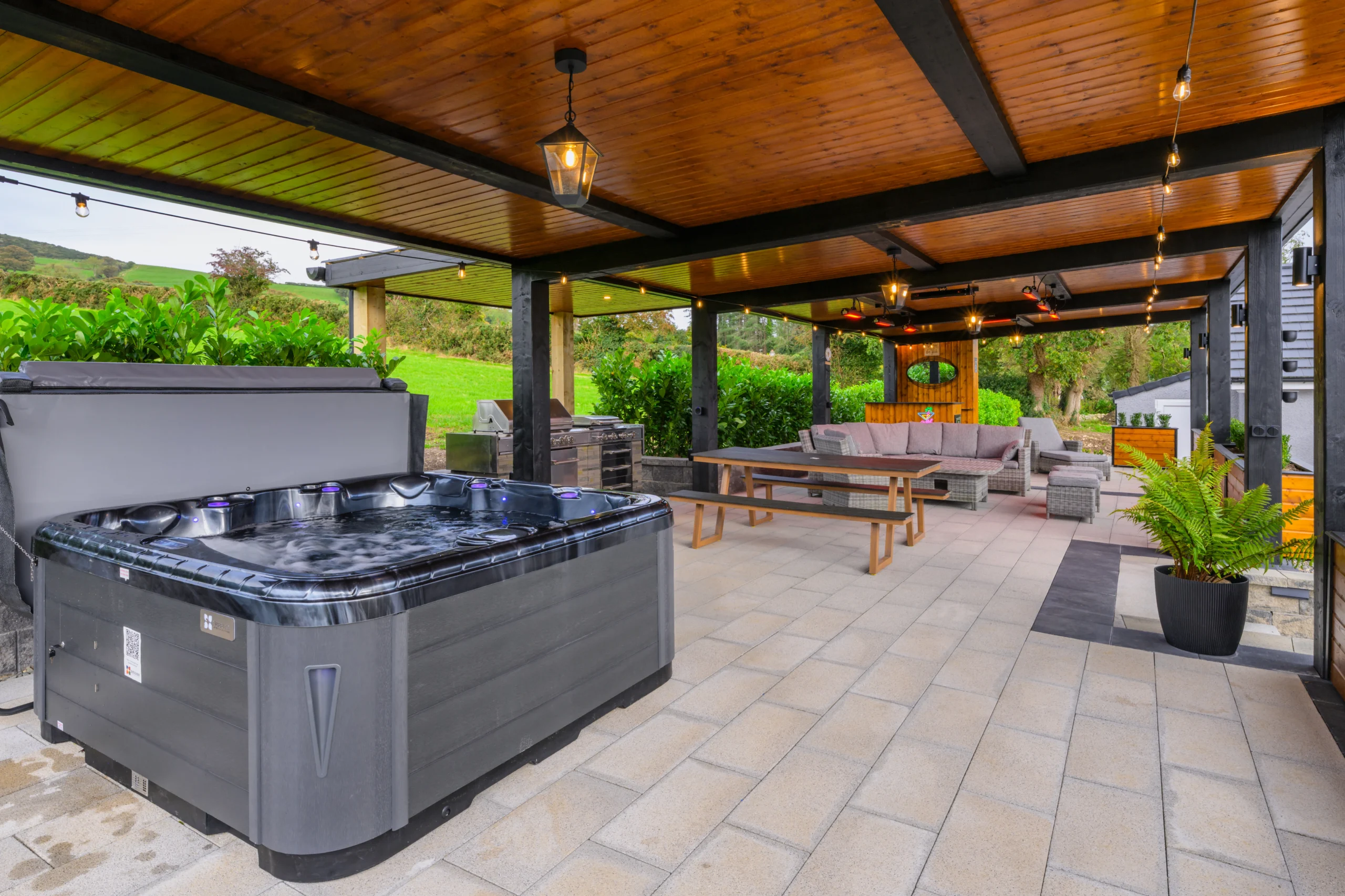 An image of the relaxation area at Rosie's Cottage, the image shows the hot tub, bench, grill, couches, and outdoor bar area.