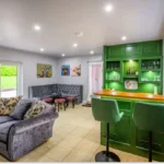 An image showcasing the green indoor bar area with various seating options, including a couch, full-size stools, small stools, and padded bench areas.