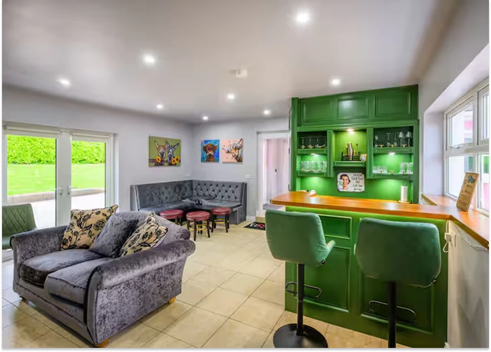 An image showcasing the green indoor bar area with various seating options, including a couch, full-size stools, small stools, and padded bench areas.