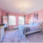 An image of a Lily's pink house bedroom decorated with pink walls, artwork, a full size mirror, two nightstands on each side of the bed, and a dresser.