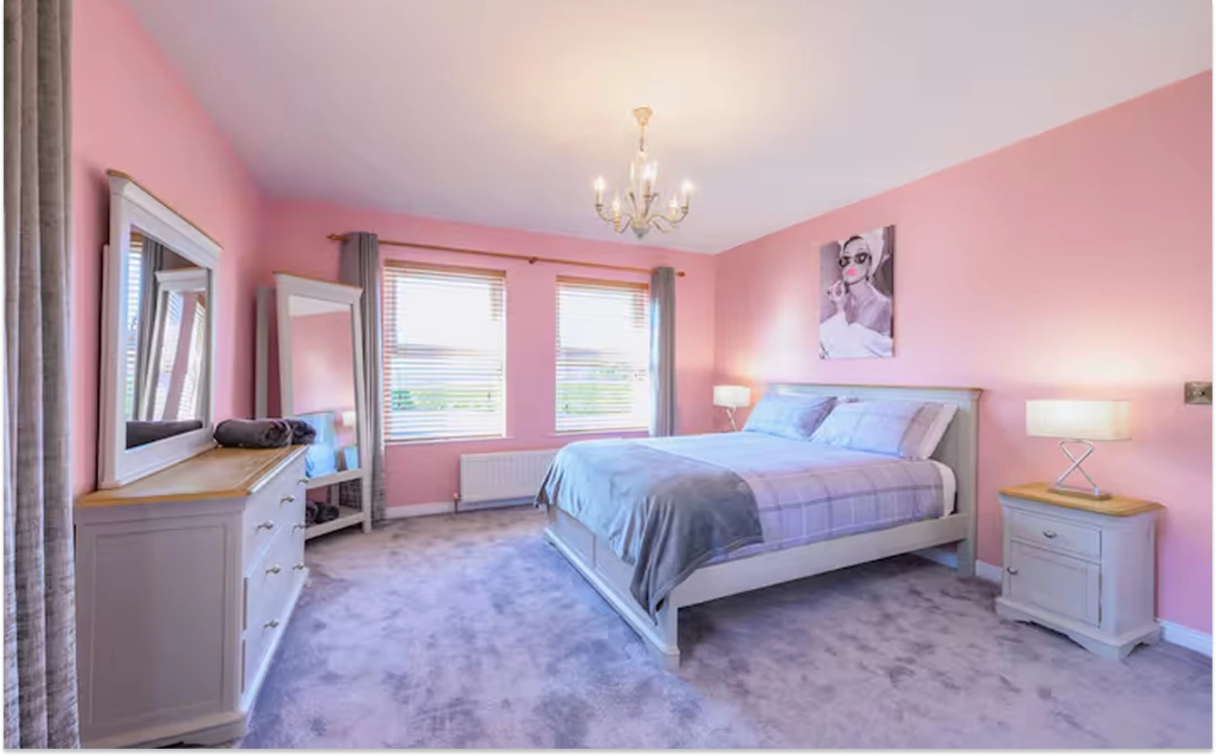 An image of a Lily's pink house bedroom decorated with pink walls, artwork, a full size mirror, two nightstands on each side of the bed, and a dresser.