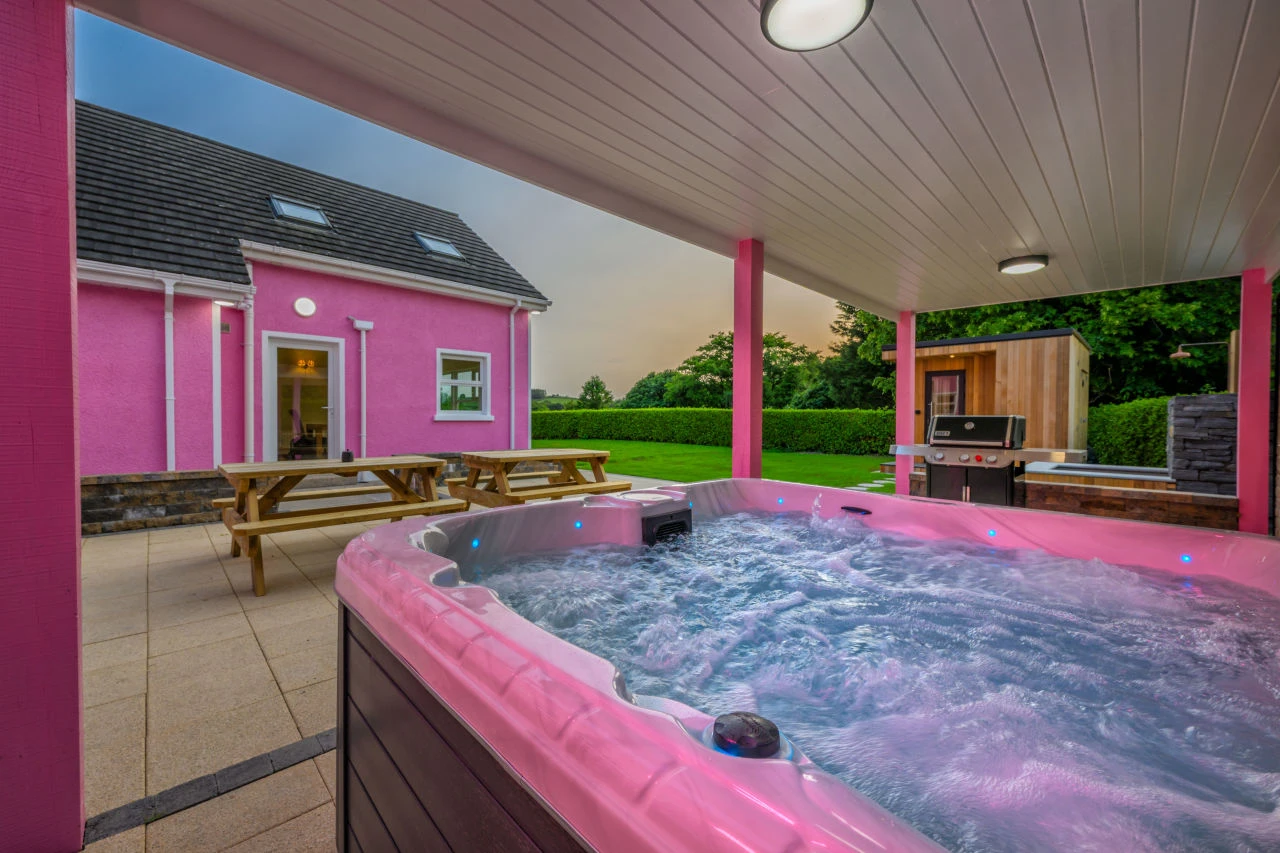 An image of the view from next to the hot tub at Lily's Pink House showcasing the sunset, back entrance into the house, grill, plunge pool, and sauna.