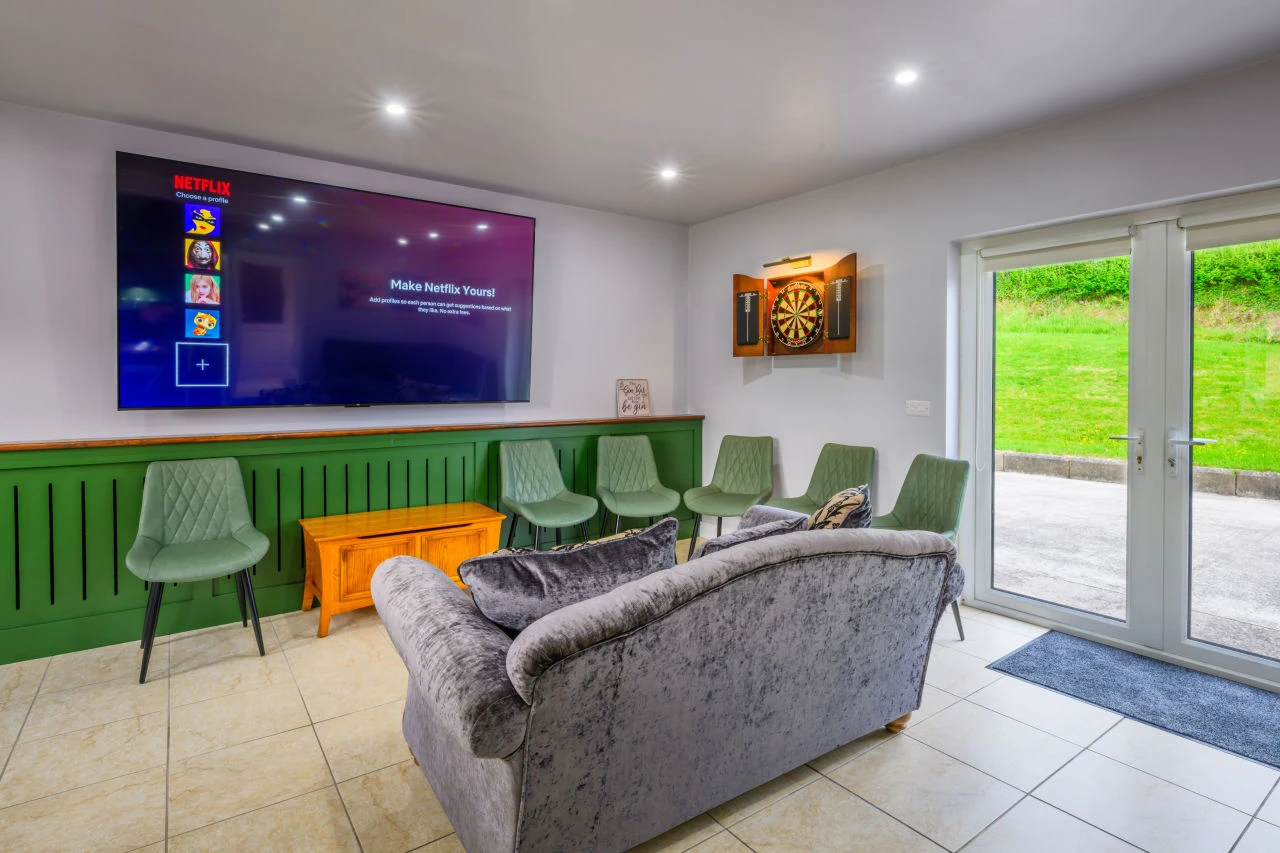 An image showcasing one of the relaxation areas in the house with a dart board, a large TV, a couch, and many different chairs around the room.