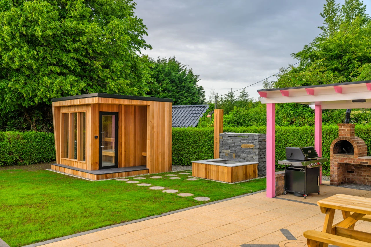 An image of Lily's Pink House outdoor grill and pizza oven area with the sauna and plunge pool in the background and the wooden bench to the side.