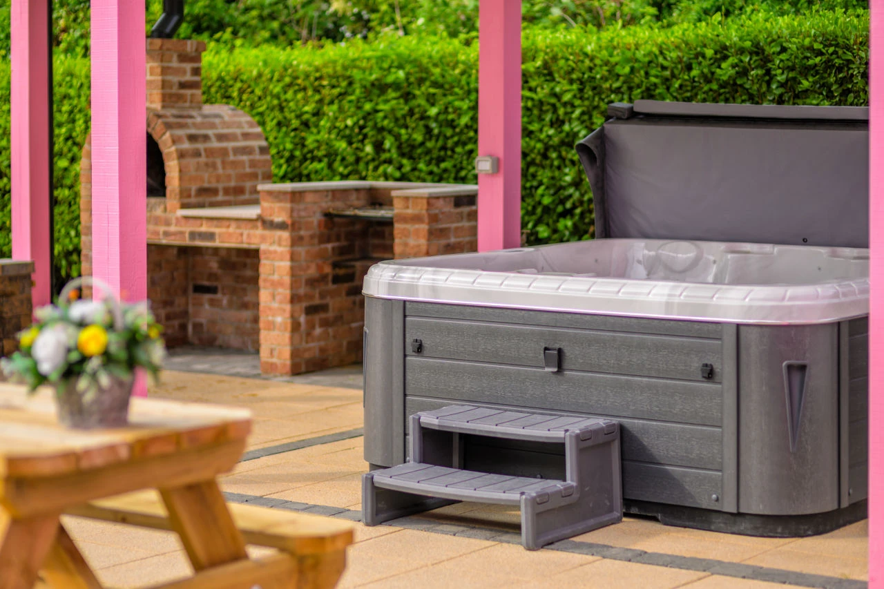 An image of Lily's Pink House hot tub with one of the benches in the foreground and the brick pizza oven in the background on the left