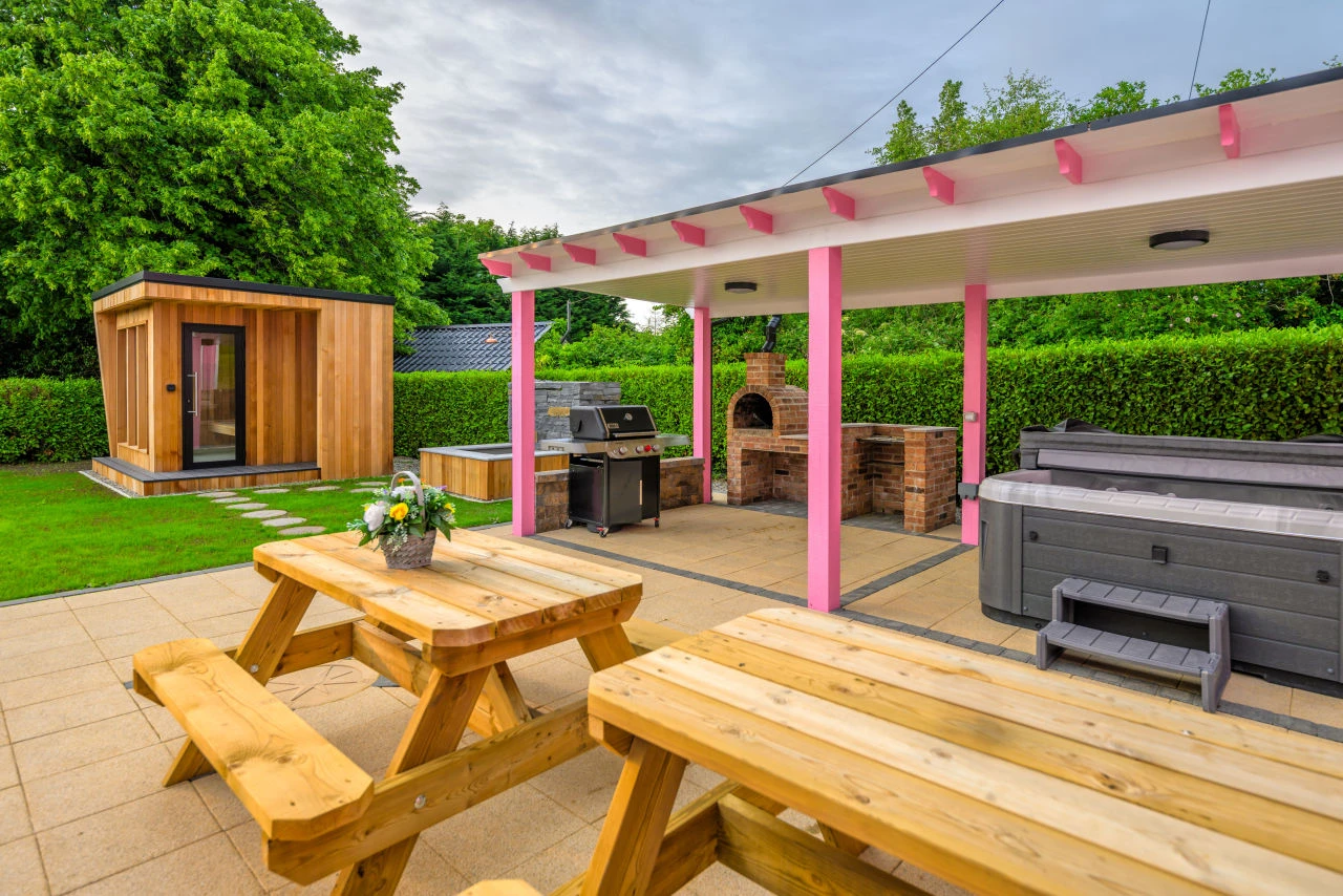 An image outside of Lily's Pink House showing the entire relaxation area with two benches in the foreground, the covered hot tub, grill, and pizza oven on the right hand side of the background