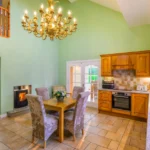 An angle of Lily's Pink house kitchen that shows the upstairs balcony overlooking the room, the living room through an open archway, a table, a fireplace, and various appliances throughout the kitchen area.