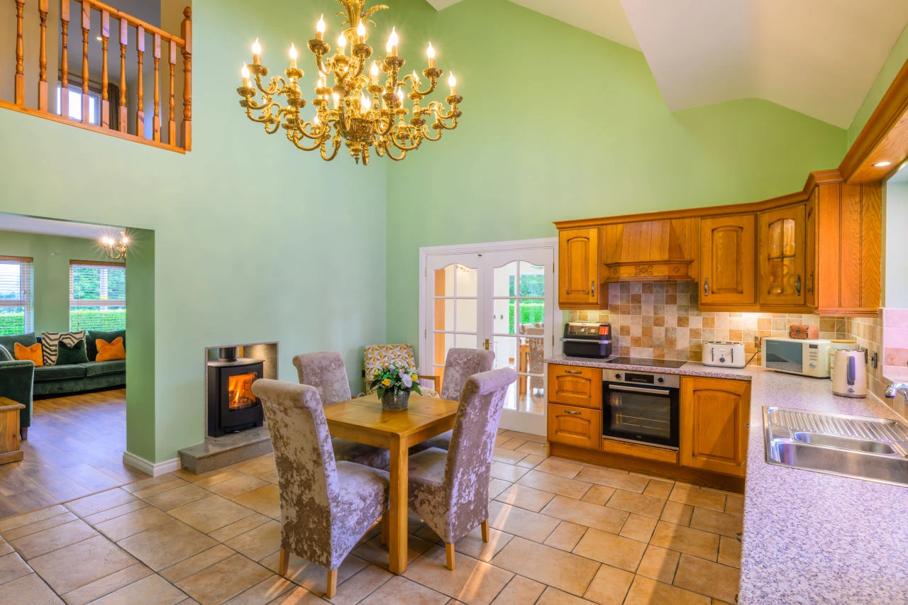 An angle of Lily's Pink house kitchen that shows the upstairs balcony overlooking the room, the living room through an open archway, a table, a fireplace, and various appliances throughout the kitchen area.