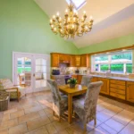 An image showing the kitchen at Lily's pink house with the fireplace, a small table, the full kitchen setup area, a golden chandelier, double doors, air fryer, coffee machine, toaster, microwave, and kettle all in view