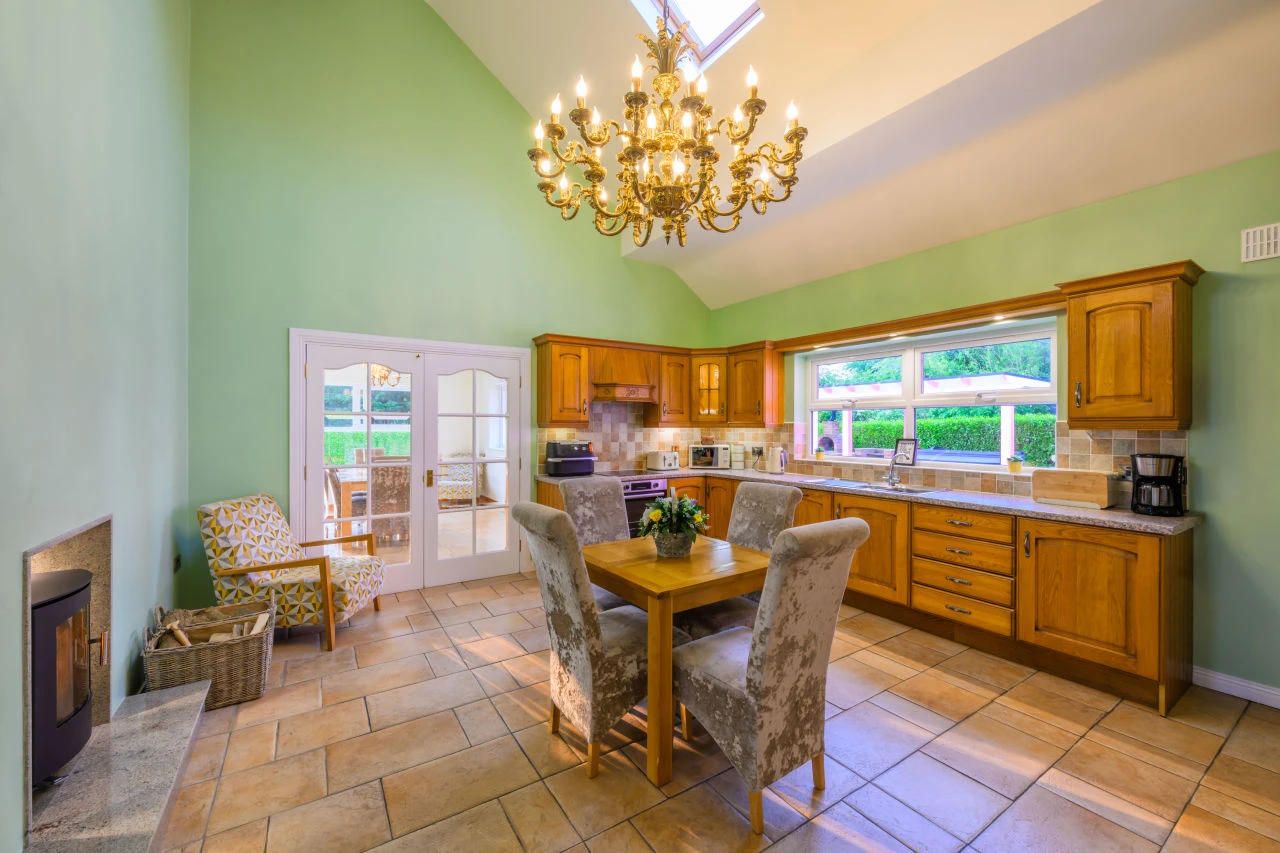 An image showing the kitchen at Lily's pink house with the fireplace, a small table, the full kitchen setup area, a golden chandelier, double doors, air fryer, coffee machine, toaster, microwave, and kettle all in view